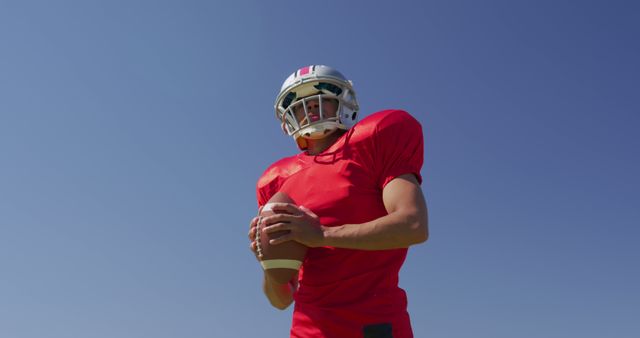 American football player wearing red jersey and helmet holds ball under clear blue sky. Ideal for use in sports-related content, promotional materials for football events, or articles about athletics.