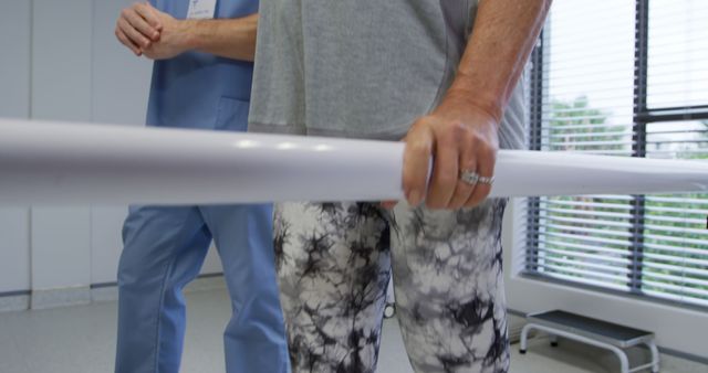 Senior patient holding onto parallel bars while a healthcare professional assists during a physical therapy session. The setting is bright and likely in a rehabilitation or medical facility, emphasizing recovery and exercise. Suitable for use in articles or advertisements focused on healthcare, recovery, senior care, physical therapy techniques, and rehabilitation programs.