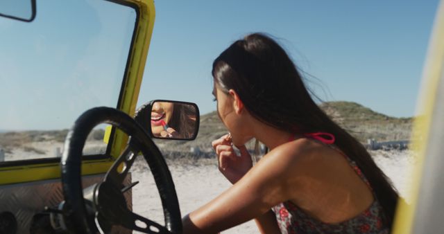 Young woman adjusting her lipstick using the rearview mirror of an off-road vehicle parked near a beach. Sunlight highlights her focused expression and the sunny, rugged outdoor background. Useful for themes related to beauty, travel, carefree lifestyle, summer adventures, and outdoor activities.