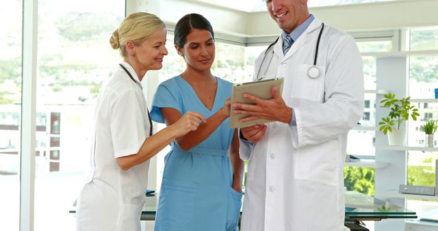 A diverse group of healthcare professionals, including a Caucasian female nurse, a Middle Eastern female doctor, and a Caucasian male doctor, are collaborating over a digital tablet, with copy space. They are engaged in a discussion, reviewing patient information or medical data in a bright hospital setting.