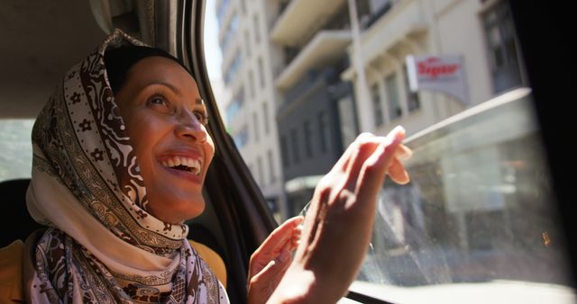 Close-up of smiling young woman in hijab looking joyfully out car window in urban setting. Ideal for depicting positive experiences of Muslim women, travel, diverse culture, urban exploration, happiness, and modern city life.