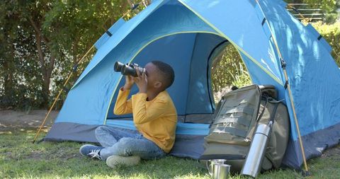 Boy exploring backyard campsite with binoculars beside blue tent and backpack in morning