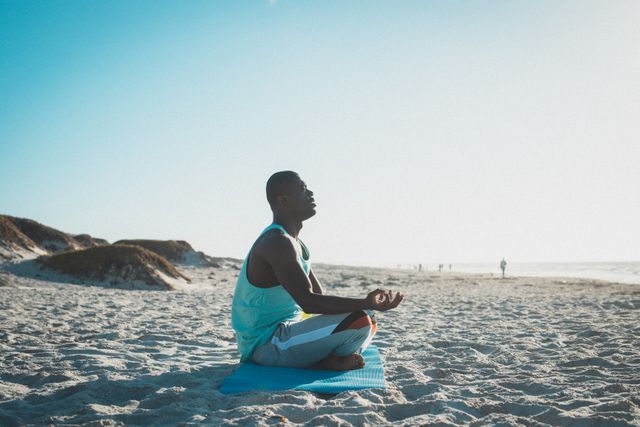 African American man practicing yoga and meditating on a beach at sunset. Ideal for promoting outdoor fitness, healthy lifestyle, mindfulness, and relaxation. Perfect for wellness blogs, fitness websites, and advertisements focusing on mental and physical health.