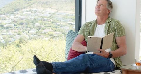 Mature man relaxing on sunlit window seat reading journal with scenic suburban view