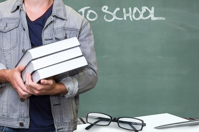 Midsection of man holding books while standing by eyeglasses on table agaisnt blackboard - Download Free Stock Photos Pikwizard.com