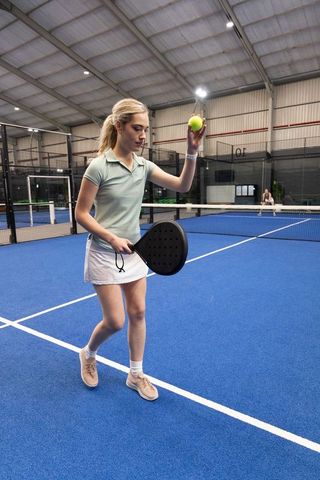 Female player serving padel ball on indoor blue court holding paddle and focusing