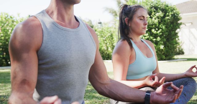 The picture shows an athletic man and woman sitting cross-legged outdoors, meditating on a sunny day. This image can be used for content related to outdoor fitness, yoga, meditation, mindfulness, health, wellness, relaxation techniques, and promoting a healthy lifestyle.