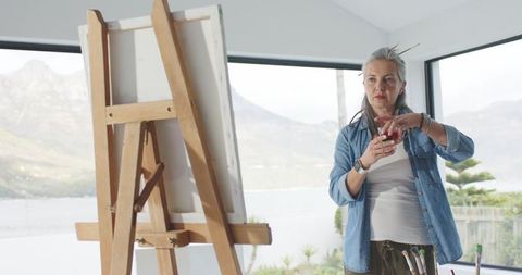 Senior woman painter standing by easel overlooking lake holding brushes and sipping drink
