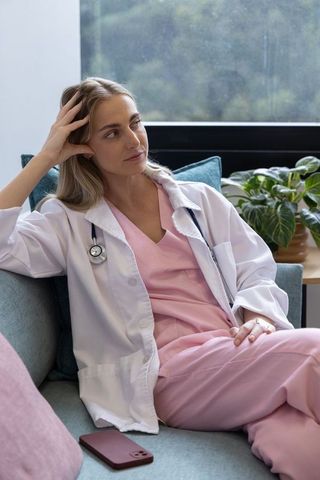 Contemplative female nurse in pink scrubs sitting on couch leaning and gazing out window