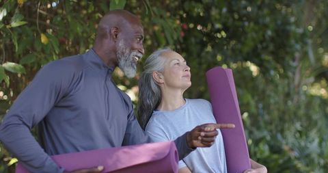 Mature couple carrying purple yoga mats in sunlit garden preparing for outdoor yoga