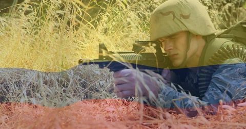 Soldier aiming assault rifle while lying prone in dry grass wearing helmet and combat gear