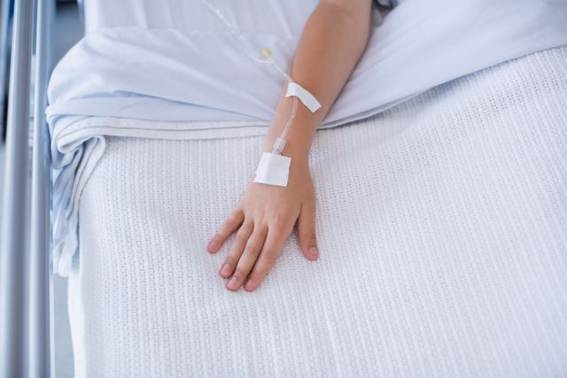 This image shows a child's hand with an IV drip on a hospital bed, representing medical treatment and healthcare. It can be used in articles or websites related to pediatric care, hospital stays, medical treatments, healthcare services, and recovery processes.