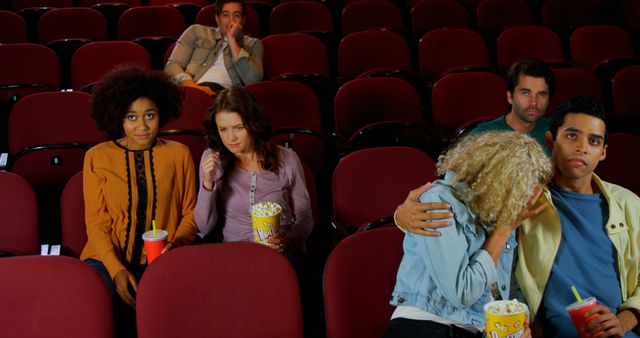 A diverse group of young adults is sitting in a movie theater, with one couple appearing to be having an emotional moment, with copy space. Their varied reactions range from surprise to discomfort, capturing a snapshot of social dynamics during a cinematic experience.