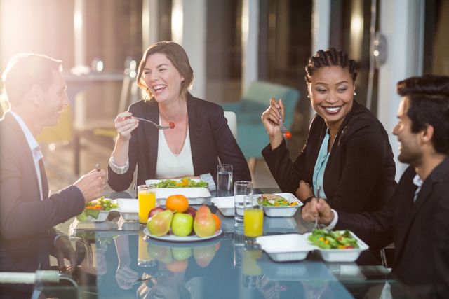 Business professionals enjoying a breakfast meeting in an office setting. They are smiling and engaging in conversation while eating healthy food. This image is ideal for illustrating corporate culture, teamwork, and healthy workplace habits in business presentations, websites, and promotional materials.