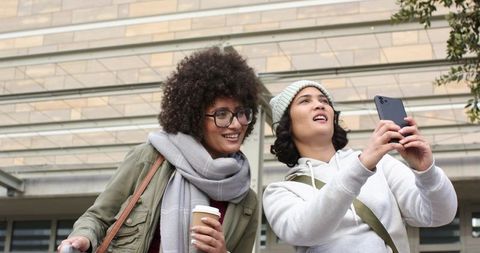 Diverse female friends taking selfie outside modern building holding coffee wearing beanie