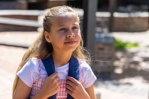 Sunlit white preteen holding backpack straps, wearing pink gingham dress, looking thoughtful