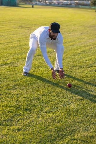Bending cricketer reaching and retrieving red ball on sunlit grass field