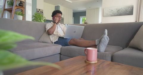 African american man working remotely on laptop reclining on grey couch and talking on phone