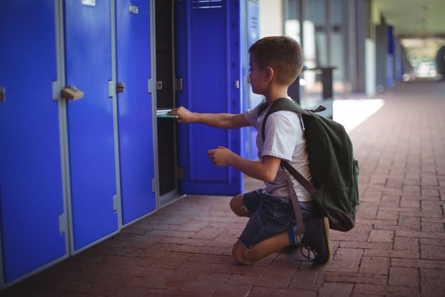 Young boy kneeling in school corridor, placing books in blue locker. Ideal for educational content, back-to-school promotions, and articles about student life and organization.
