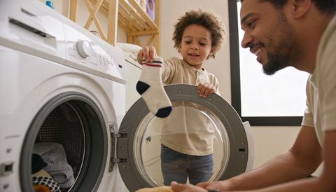 Father teaching young boy sorting socks at front-loading washer, smiling bonding moment