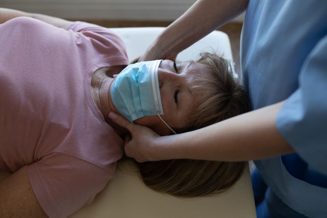Senior Caucasian woman at home visited by Caucasian female nurse, massaging her neck, wearing face masks. Medical care
at home during Covid 19 Coronavirus quarantine.
