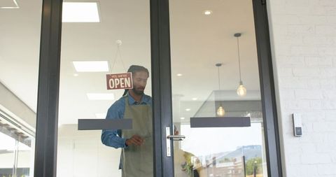 African american small business owner opening glass door with hanging open sign and apron