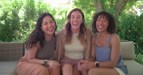 Diverse friends laughing on backyard sofa wearing smartwatches and casual summer outfits