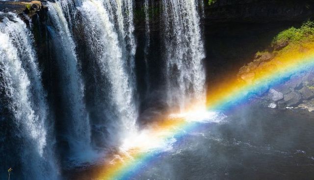 Cascading waterfall creating vibrant rainbow arc across sunlit gorge