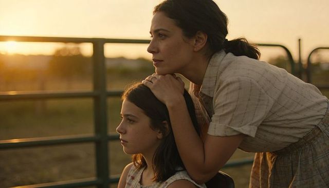 Leaning mother supporting daughter by ranch gate during golden hour, tender family moment