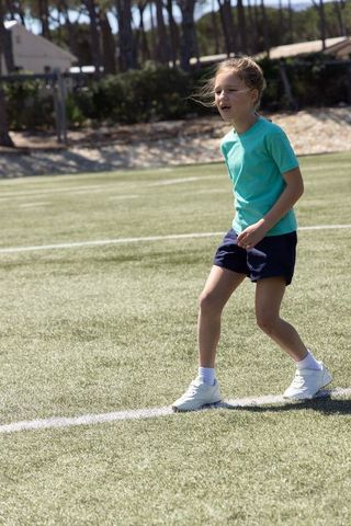 Young girl walking on soccer field in teal shirt and white sneakers, sunny outdoor activity