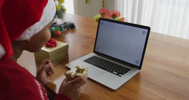 Child wearing Santa hat enjoying a holiday celebration via laptop. Christmas decorations and a wrapped gift are on the table. Perfect for themes about virtual celebration, technology in holidays, and festive family moments.