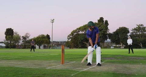 Cricket batsman swinging bat at twilight on suburban field wearing green helmet and pads