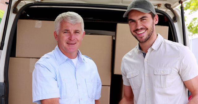 Two male delivery workers stand smiling next to a vehicle filled with boxes. They are outdoors and appear professional in their uniforms, making them ideal for illustrating concepts related to shipping, logistics, transportation services, and moving businesses.