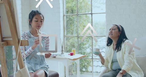 Two women painting together at easel in sunlit studio with large window and greenery