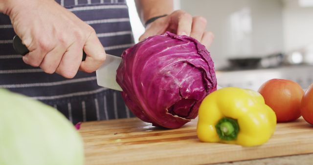 This image showcases a person slicing a red cabbage on a wooden cutting board with a knife. Nearby, a yellow bell pepper and tomatoes are present, suggesting a focus on fresh ingredients and home cooking. Ideal for culinary blogs, healthy eating promotions, vegetarian recipe books, cooking classes, or lifestyle articles about home cooking and kitchen activities.