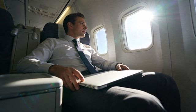Executive traveling with laptop in premium airplane window seat bathed in warm sunlight