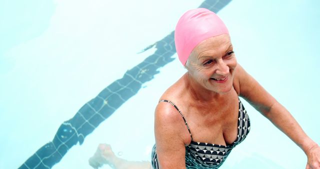 A senior Caucasian woman enjoys her time in a swimming pool, smiling as she engages in physical activity. Her pink swim cap and patterned swimsuit suggest a lively spirit and dedication to fitness.