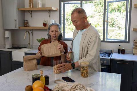 Asian couple pouring chickpeas into jar while organizing pantry on sunlit kitchen island