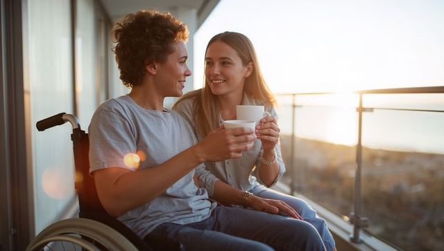Couple sipping coffee on balcony at sunset, manual wheelchair visible, warm urban moment