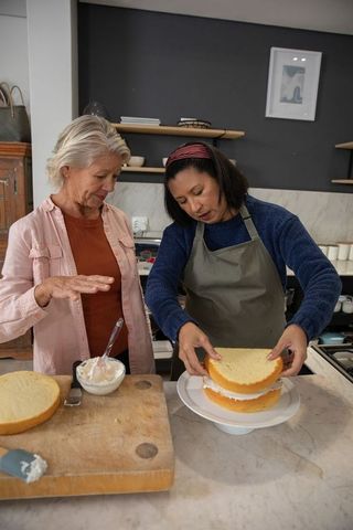 Senior women assembling layered sponge cake together in cozy home kitchen teaching baking