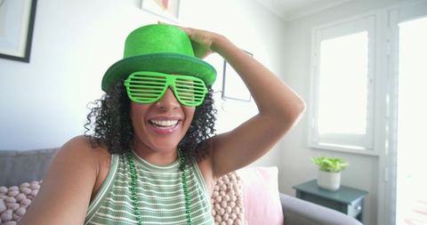 African american woman celebrating at home wearing green hat and shutter shades, smiling