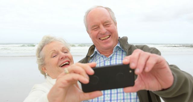 This stock photo showcases a cheerful senior couple taking a selfie on a beach, perfect for promoting retirement, vacation destinations, healthy aging, lifestyle content, senior relationships, and marketing campaigns targeting older adults enjoying life to the fullest.