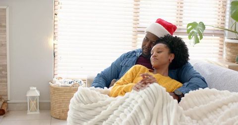 Cozy diverse couple cuddling on sofa under plush white blanket wearing santa hat