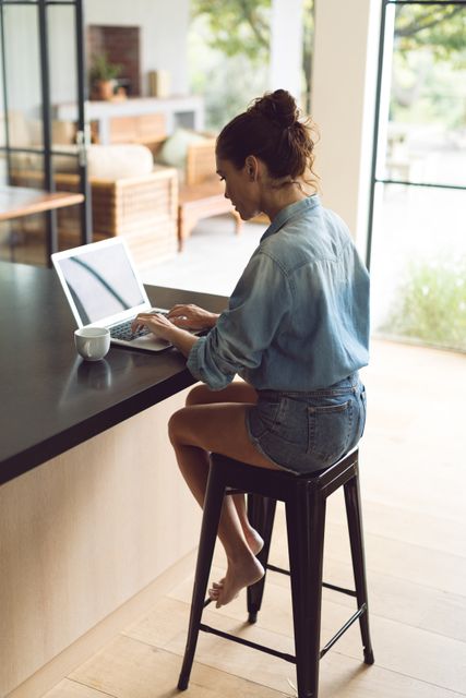 Woman sitting on stool at kitchen counter, using laptop. She is wearing casual denim outfit. Bright, modern kitchen with natural light. Ideal for concepts of remote work, productivity, modern lifestyle, and home office.