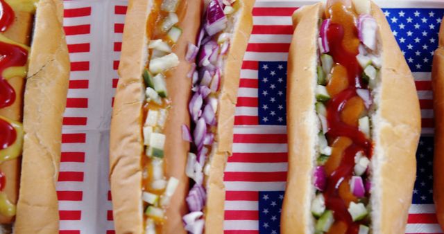 Hot dogs arranged on American flag background feature various toppings, including onions, pickles, mustard, and ketchup. Perfect for celebrating national holidays, picnics, barbecues, or summer gatherings. Vibrant ingredients add a festive note, making it fitting for marketing materials, advertisements, or social media campaigns related to American traditions and cuisine.