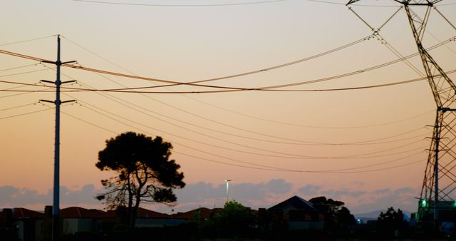 Silhouetted electric power lines stretch above suburban buildings during sunset. Orange and pink hues fill the sky, creating a serene evening atmosphere. Useful for energy sector themes, urban landscape backgrounds, or depicting suburban living at dusk.