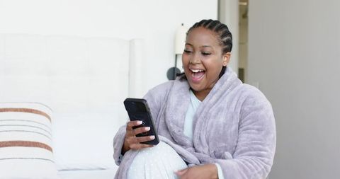 Midlife african american woman relaxing on bed in lavender robe using smartphone, smiling