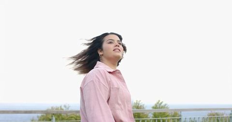 Young woman standing seaside railing gazing toward horizon windswept hair pastel pullover