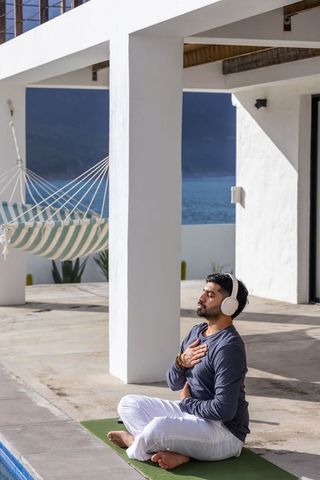 Coastal mindfulness session: indian man meditating on terrace by pool with headphones