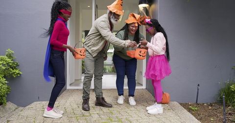 Diverse family handing out Halloween candy on porch wearing orange hats and pumpkin buckets